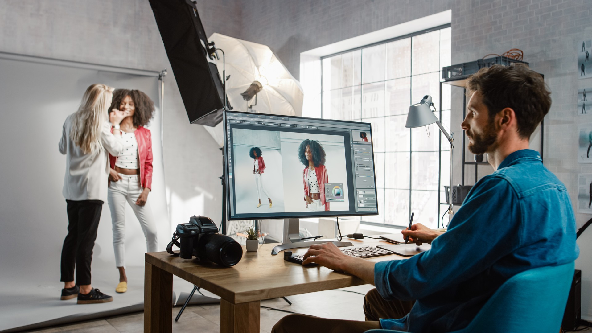 Backstage of the Photoshoot: Make-up Artist Applies Makeup on Beautiful Black Girl. Photo Editor Works on Desktop Computer Retouching Photo with Image Editing Software. Fashion Magazine